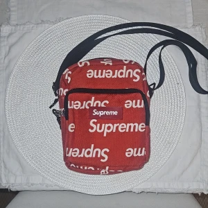 Röd axelväska från Supreme - Säljer en cool röd axelväska från Supreme med deras ikoniska logga i vitt. Väskan har en justerbar axelrem och en praktisk dragkedja framtill. Perfekt för att bära med sig det viktigaste på ett stilfullt sätt.