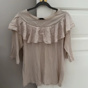 Beigerosa topp med volang från Lindex - Säljer en söt beige topp från Lindex med volangdetaljer runt halsen. Toppen har trekvartsärmar och en avslappnad passform. 