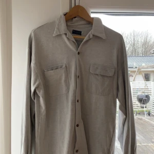 Grå overshirt från Bondelid - Snygg grå långärmad skjorta från Bondelid med klassisk krage och knappar framtill. Skjortan har två bröstfickor med lock och broderad logga på ena fickan. Perfekt för en stilren look. Storlek L men passar bra på M
