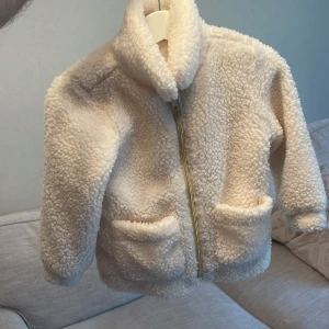 Beige teddyjacka från H&M - Mysig beige teddyjacka från H&M för de små. Jackan har en dragkedja framtill och två praktiska fickor. Perfekt för kyliga dagar med sin fluffiga och varma design. 🧸