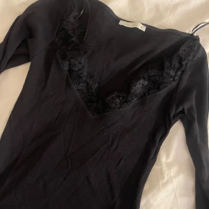 Svart ribbad topp från Zara - Säljer en stilren svart ribbad topp från Zara med spetsdetaljer vid halsringningen. Toppen har långa ärmar och en v-ringad design som ger en elegant touch. Perfekt för att klä upp eller ner beroende på tillfälle.