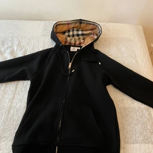 Svart hoodie från Burberry - Säljer en stilren svart hoodie från Burberry med dragkedja och klassiskt rutigt foder i huvan. Perfekt för en avslappnad och trendig look. Huvan har justerbara snören och jackan har långa ärmar.
