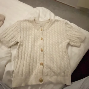 Vit stickad kofta från H&M - Säljer en söt vit stickad kofta från H&M med korta ärmar och fina guldfärgade knappar.  Väldigt bra skick då jag använt den ett få tal gånger då den inte kommit till användning 🩷.