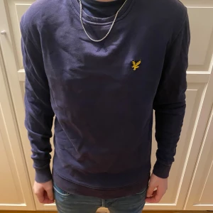 Mörkblå tröja med broderad logga - Säljer en stilren mörkblå tröja från Lyle & Scott med deras ikoniska broderade logga på bröstet. Tröjan har långa ärmar och en rund halsringning. Perfekt för en avslappnad look.