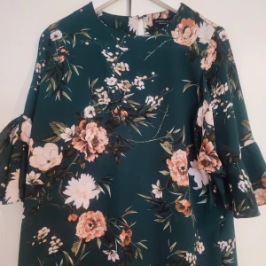 Blommig blus från Dorothy Perkins - Säljer en vacker blommig blus från Dorothy Perkins i grönt med rosa och vita blommor. Klänningen har trekvartsärmar med volangdetaljer och en rund halsringning. Perfekt för en somrig look! 🌸