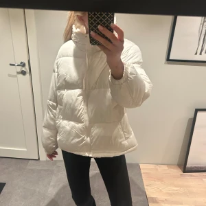 Vit pufferjacka från H&M - Säljer en snygg vit pufferjacka från H&M. Jackan har en dragkedja framtill och en mysig huva med snörning. Perfekt för kyliga dagar med sin vadderade design och långa ärmar.