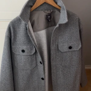 Grå overshirt från H&M - Säljer en stilren grå overshirt från H&M. Jackan har en klassisk krage och knäppning framtill med svarta knappar. Två stora bröstfickor med lock och knappar ger en cool look. Perfekt för lager-på-lager-stil.