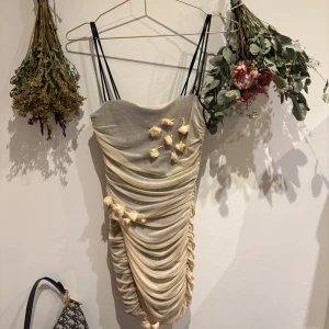 Zara summer dress - Säljer en elegant beige klänning med rynkdetaljer och axelbandslös design. Klänningen har dekorativa blommor framtill och smala axelband. Perfekt för en stilfull kväll!