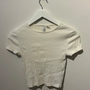 Vit ribbad topp från H&M  - Säljer en stilren vit ribbad topp från H&M Divided. Den har en klassisk rund hals och korta ärmar, perfekt för en enkel och snygg look. Passar bra till både jeans och kjol.