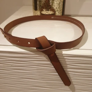 Brunt skärp i skinnimitation - Snyggt brunt skärp i skinnimitation. Det har en enkel design med flera hål för justering och en stilren knutdetalj framtill. Perfekt för att ge en elegant touch till din outfit.