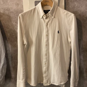 Vit långärmad skjorta från Ralph Lauren - Säljer en stilren vit långärmad skjorta från Ralph Lauren. Skjortan har en klassisk krage och knappar framtill. Den är broderad med den ikoniska Ralph Lauren-loggan i mörkblått på bröstet. Perfekt för en elegant look! Storlek Small och i väldigt bra skick!