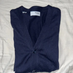 Mörkblå kofta från Selected Homme - Säljer en stilren mörkblå kofta från Selected Homme. Koftan har en v-ringad design med knappar framtill och är tillverkad i ett mjukt material. Perfekt för lager-på-lager under kyligare dagar.