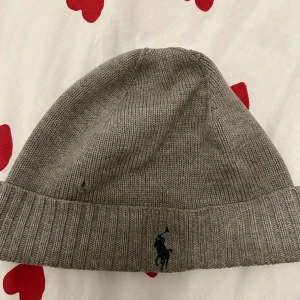 Grå stickad mössa från Polo Ralph Lauren - Säljer en stilren grå stickad mössa från Polo Ralph Lauren. Mössan har en ribbad kant och är dekorerad med det ikoniska broderade logotypen i svart. Perfekt för kyliga dagar. Ett litet hål till vänster 