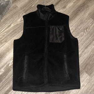 Svart fleeceväst med dragkedja - Snygg svart fleeceväst med dragkedja och en bröstficka i ett glansigt material med storlek S. Perfekt för lager-på-lager under kyliga dagar. Västen har en stilren design med två sidofickor.