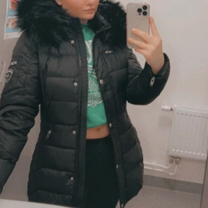 Svart dunjacka med pälsdetaljer - Snygg svart dunjacka med fluffig fusk päls på huvan. Jackan har dragkedja och knappar framtill samt praktiska fickor med dragkedja. Perfekt för kalla dagar. Dock finns det ett litet hål i mitten av ryggen. Inget man lägger märke till. 