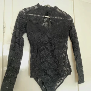 Svart spetsbody från Bik Bok - Säljer en elegant svart spetsbody från Bik Bok. Bodyn har långa ärmar och en hög hals med en fin spetsdesign. Perfekt för att ge en stilfull touch till din outfit.