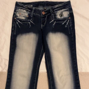 Mörkblå jeans med broderade detaljer - Snygga mörkblå jeans med unika broderade detaljer på bakfickorna och en cool tvättad look. Byxorna har en knapp och dragkedja framtill samt dekorativa sömmar. Perfekta för dig som vill sticka ut med stil!