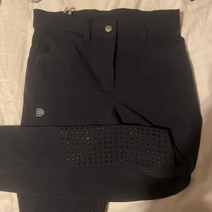 Mörkblå ridbyxor med glitterdetaljer - Snygga mörkblå ridbyxor med glittrande detaljer och silikonförstärkning vid knäna. Byxorna har en knapp och dragkedja framtill samt en glittrig detalj på bakfickan. Perfekta för unga ryttare som vill ha stil och funktion.        Köpte dem för 899kr där av säljer jag dem för 450kr