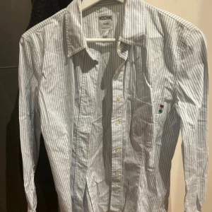 Randig skjorta från Moschino - Snygg figursydd randig skjorta från Moschino i blått och vitt. Skjortan har långa ärmar och knappar framtill. Perfekt för en stilren look med en liten logga på bröstfickan. Tillverkad i Italien. Storlek M. Stretchigt tyg. 96% bomull 4% elsatsningar