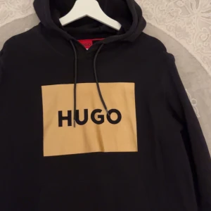 Svart hoodie från Hugo - Säljer en stilren svart hoodie från Hugo med en beige rektangel och svart logotyp på bröstet. Hoodien har en justerbar dragsko i huvan och är perfekt för en avslappnad look. Passar bra till både jeans och joggers. (Köpt för 899)