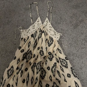 Mönstrad klänning med spetsdetaljer - Säljer en snygg klänning med svart mönster på beige bakgrund. Klänningen har smala axelband och fina spetsdetaljer upptill. Perfekt för en somrig look!