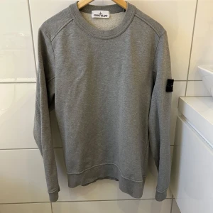 Stone Island - Säljes: En original Stone Island sweatshirt i klassisk Grå. Denna tröja är det perfekta valet för dig som vill ha en stilren och tidlös look med en touch av lyx.  Detaljer: • Marke: Stone Island • Färg: grå • Storlek: M • Skick: 10/10 • Material: Bekvämt och slitstarkt tyg  Tröjans ikoniska Stone Island-kompasslogotyp på vänster ärm gör den igenkännbar och ger en exklusiv touch. Den är perfekt för bäde vardagsbruk och vardagliga tillfällen.