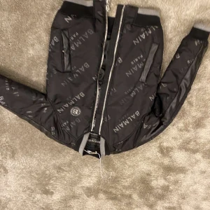 Svart bomberjacka från Balmain - Snygg svart bomberjacka från Balmain med logotyptryck över hela jackan. Den har en dragkedja framtill och ribbade muddar vid ärmslut och nederkant. Perfekt för en stilren look.