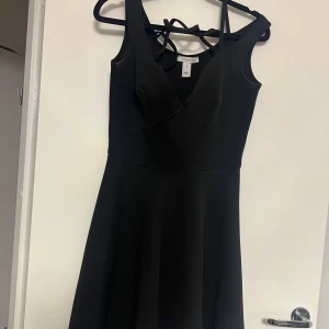 Svart klänning med omlott-design - Elegant svart klänning med omlott-design och djup v-ringning. Klänningen har en stilren och tidlös look, perfekt för olika tillfällen. Den är ärmlös och har en smickrande passform som framhäver figuren.