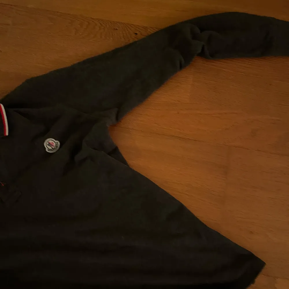 Säljer en stilren grå pikétröja från Moncler med klassisk krage och knappar. Tröjan har en broderad logotyp på bröstet och röd-vit randig detalj på kragen. Perfekt för en avslappnad men ändå elegant look. Du kan bekräfta autenticitet via QR kod på sista bilden .. Neuletakit & Villapaidat.
