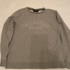 Grå tröja från Sail Racing - Säljer en stilren grå tröja från Sail Racing med texten 'SAIL RACING EST. 1977' på framsidan. Tröjan har långa ärmar och en rund halsringning. Perfekt för en avslappnad stil.