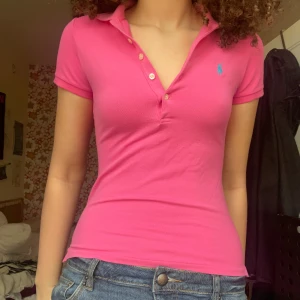 Rosa pikétröja från Ralph Lauren - Säljer en snygg rosa pikétröja från Ralph Lauren med korta ärmar och klassisk krage. Tröjan har knappar framtill och en liten blå logga på bröstet.