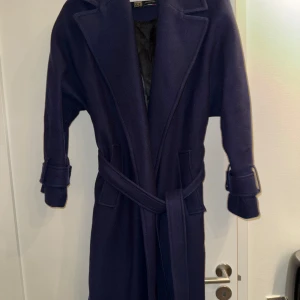 Mörkblå kappa från Zara manteco ull (75%) storlek S - Elegant mörkblå kappa från Zara med bälte i midjan och knappar vid ärmsluten. Tillverkad av högkvalitativt tyg från Manteco. Perfekt för kyligare dagar med sin långa design och klassiska stil. Storlek S.