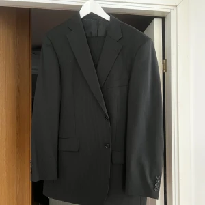 Svart randig kavaj från Elson - Snygg svart kostym från Elson i ull med diskreta ränder. Kavajen har två knappar framtill och klassiska fickor. Perfekt för en stilren look.