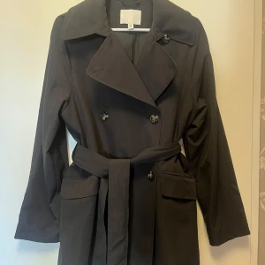 Svart trenchcoat med bälte - Snygg svart trenchcoat med knappar och bälte i midjan. Jackan har långa ärmar och en klassisk krage. Perfekt för en stilren look. Storlek xs, men oversize i modellen så tycker egentligen att det är en M/S. 
