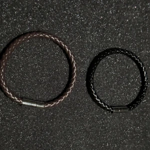 Två pack flätade armband i läder - Säljer två stilrena flätade armband i veganskt läder, ett i brunt och ett i svart. Båda har en metallisk låsning som ger en modern touch. Perfekta för att komplettera din outfit med en elegant detalj.