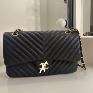 Svart quiltad axelväska med kedja - Elegant svart axelväska i quiltat mönster med guldfärgad kedja och detaljer. Väskan har en praktisk ficka på baksidan och stängs med en CC tryckknapp. Perfekt för att ge en stilren touch till din outfit.