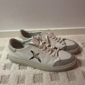 Vita sneakers med fågelbroderi - Snygga vita sneakers från Axel Arigato med ett coolt fågelbroderi på sidan. Skorna har grå mockadetaljer och klassisk snörning. Perfekta för en stilren look.