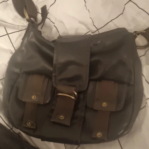 Brun axelväska med fickor - Säljer en snygg brun axelväska med praktiska fickor framtill och ett spänne. Väskan har en justerbar axelrem och stängs med dragkedja. Perfekt för att bära med sig det nödvändigaste.