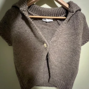 Beige stickad kofta från Zara - Säljer en mysig beige stickad kofta från Zara. Koftan har korta ärmar och knappar framtill. Perfekt för lager-på-lager under kyligare dagar. Den har en ribbad krage och nederkant som ger en stilren look.