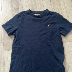 Mörkblå t-shirt från Lyle & Scott - Säljer en stilren mörkblå t-shirt från Lyle & Scott med korta ärmar. T-shirten har en gul logotyp broderad på bröstet. Perfekt för en avslappnad stil.