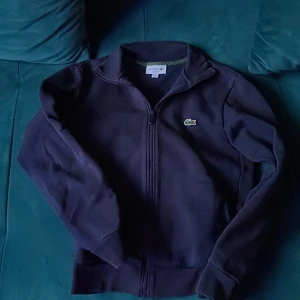 Mörkblå tröja från Lacoste - Säljer en snygg mörkblå tröja från Lacoste med dragkedja och det klassiska krokodilmärket på bröstet.  Tröjan är knappast använd pågrund av att den var för liten. 