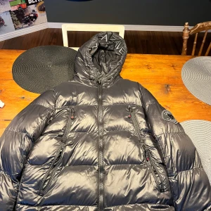 Svart dunjacka från Zavetti Canada - Säljer en svart dunjacka från Zavetti Canada med huva och dragkedja. Jackan har ett quiltat mönster och märkesdetaljer på insidan och utsidan. Perfekt för kalla dagar. Sitter perfekt på mig som är 180cm och väger 70kg