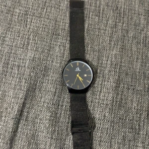 Svart klocka från Calvin Klein - Snygg svart klocka från Calvin Klein med minimalistisk design. Klockan har en svart urtavla med guldiga visare och markeringar. Armbandet är i svart meshmaterial vilket ger en modern och stilren look.