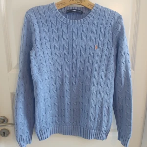 Ljusblå kabelstickad tröja från Ralph Lauren - Så snygg kabelstickad tröja från Ralph Lauren, ljust blålila med peachfärgad logga. Stl M, upplevs true to size, se mått. Mycket fint skick, inga hål eller fläckar.  Mått: Armhåla till armhåla 44,5cm, bredd längst ner 38cm, längd från mitt på axeln 62cm.