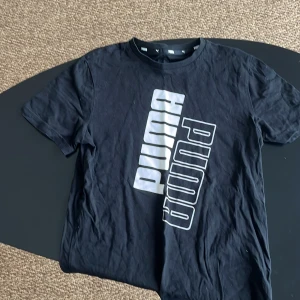 Svart t-shirt från Puma - Snygg svart t-shirt från Puma med ett stort, diagonalt tryck i vitt och grått på framsidan. Perfekt för en avslappnad stil. Tillverkad i mjukt bomullsmaterial för extra komfort.