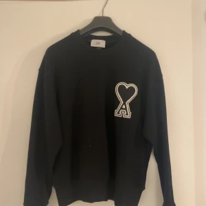 Svart Ami tröja  - Säljer en stilren svart tröja från Ami med en unik hjärtdetalj på bröstet. Tröjan har långa ärmar och en klassisk passform. Perfekt för en avslappnad look.