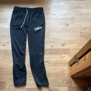 Svarta träningsbyxor från Zone Floorball - Snygga svarta träningsbyxor från Zone Floorball med vit logga på benet. Byxorna har elastisk midja med snörning och praktiska dragkedjefickor. Perfekta för innebandyträning eller annan sportaktivitet. XS 