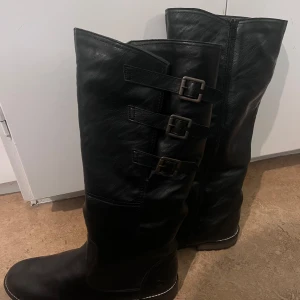 Svarta stövlar med spännen - Snygga svarta stövlar i skinn med tre dekorativa spännen på sidan. Perfekta för höst och vinter med en stilren design och dragkedja för enkel påtagning. Använda bara ett par gånger. Märkesskorna köptes för 1700kr