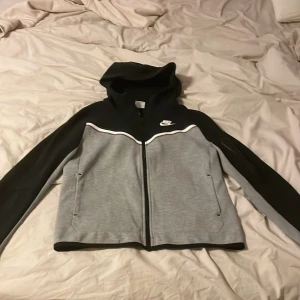 Grå och svart huvtröja från Nike - Säljer en grå och svart huvtröja från Nike med dragkedja och vit logga på bröstet. Tröjan har en huva och långa ärmar med en stilren design. Perfekt för en sportig look eller avslappnade dagar.