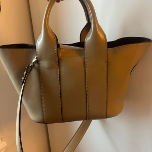 Beige handväska med axelrem - Snygg beige handväska i konst-skinn med justerbar axelrem och två handtag. Väskan har en stilren design med synliga sömmar och metallspännen. Perfekt för att bära dina dagliga nödvändigheter med stil.